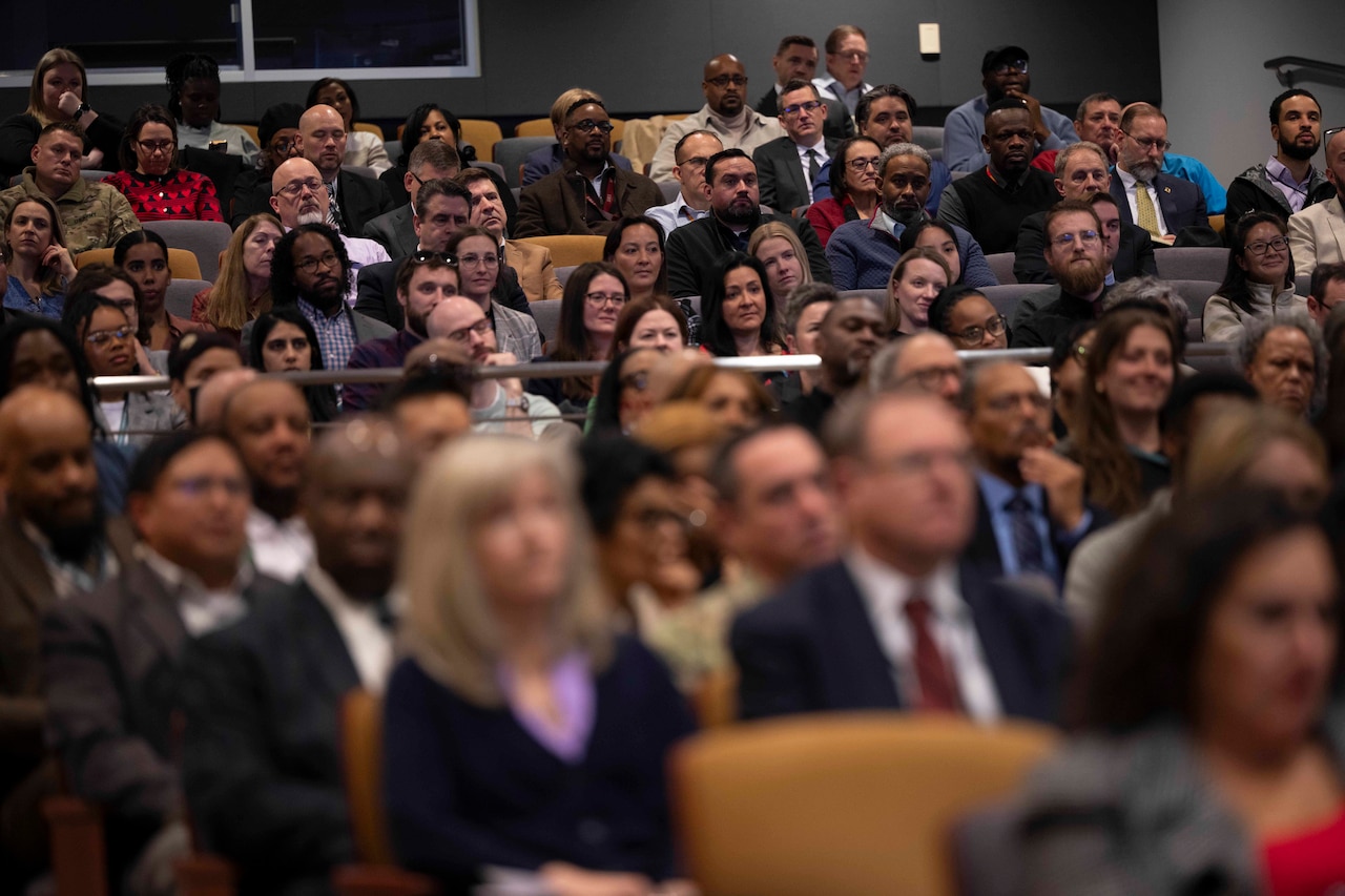 A large crowd of people in business attire seated in an auditorium observing someone speaking to them from off-screen.