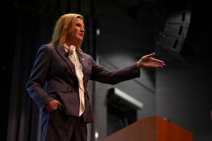 A woman in business attire stands on a stage, gesturing with her left hand while speaking to an audience off-screen.