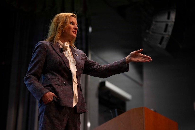 A woman in business attire stands on a stage, gesturing with her left hand while speaking to an audience off-screen.