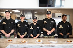A group of uniformed personnel stand side by side in front of plates of food.