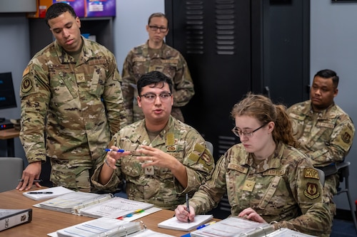 A group of men and women, wearing camouflage military uniforms, sit around a table covered in binders, maps, and documents.