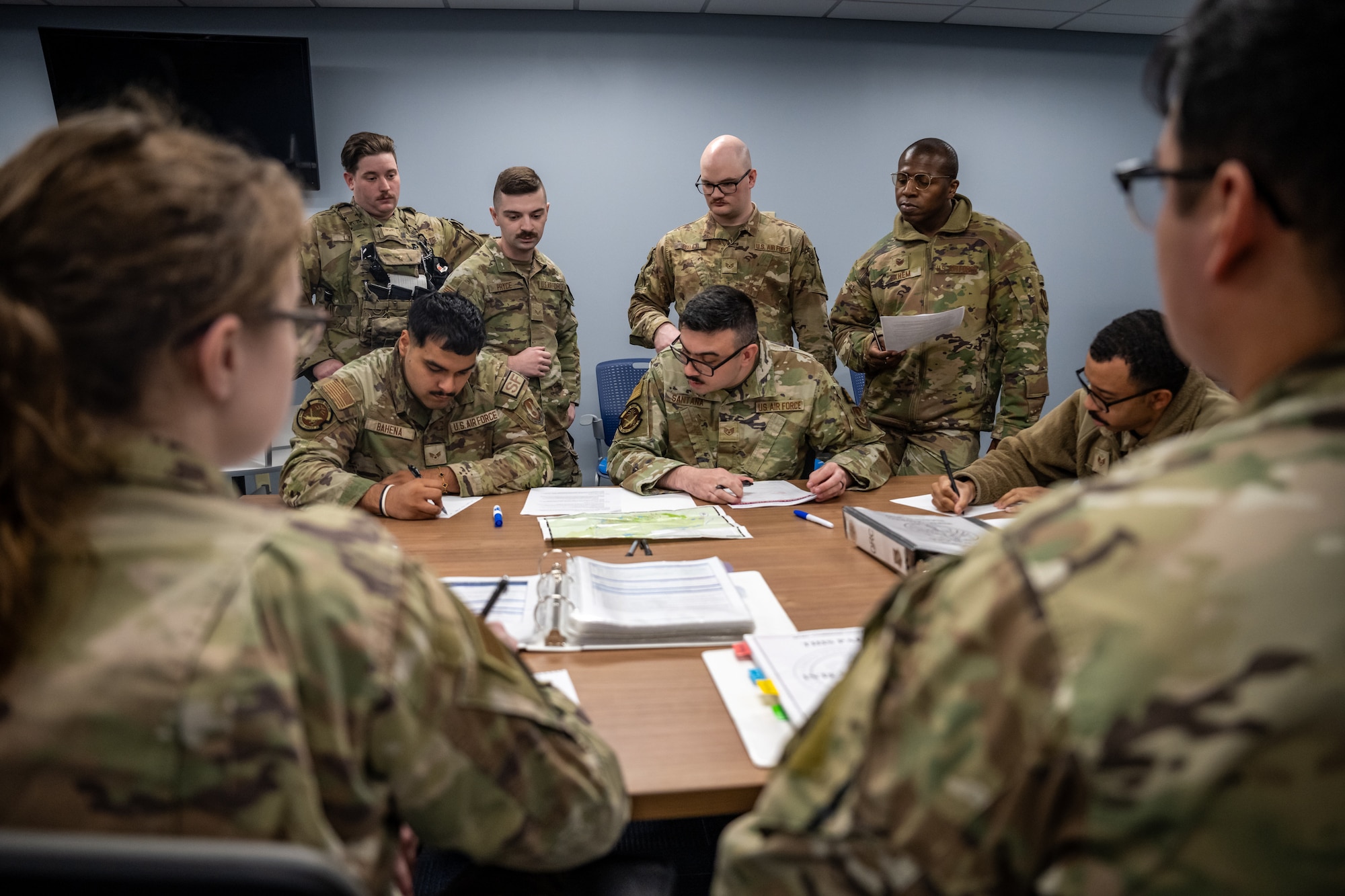 A group of men and women, wearing camouflage military uniforms, sit around a table covered in binders, maps, and documents.