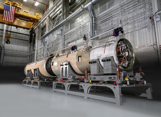 A large, cylindrical missile test booster undergoing assembly on heavy-duty stands inside a hangar, featuring exposed internal wiring at one end and an American flag suspended in the background.