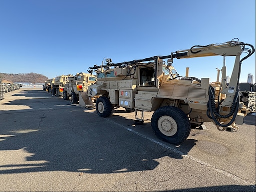 Several lines of large military vehicles are parked on a concrete tarmac.