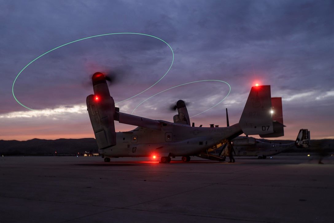 A helicopter with green glowing lights swirling around its propellers sits on a flight line under a gray, cloudy sky.