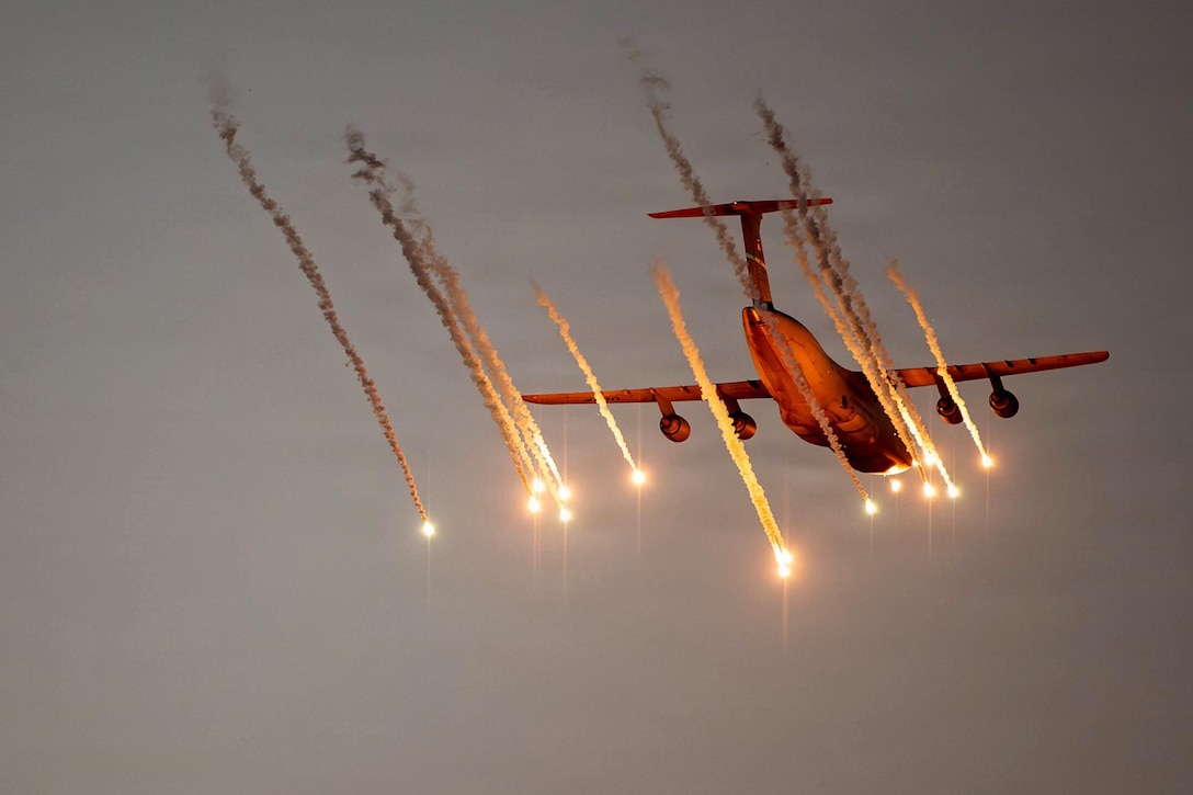A military aircraft flies through a gray, cloudy sky while white and orange flares trail behind it.