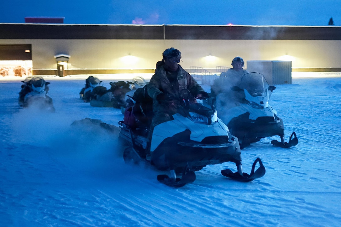 Two people wearing camouflage military uniforms, gloves and helmets ride snowmobiles, with several other snowmobiles, equipment and a building in the background.
