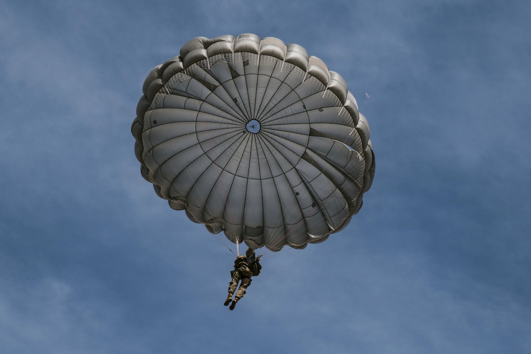 A person wearing a camouflage military uniform and helmet attached to an open parachute falls in a cloudy blue sky.