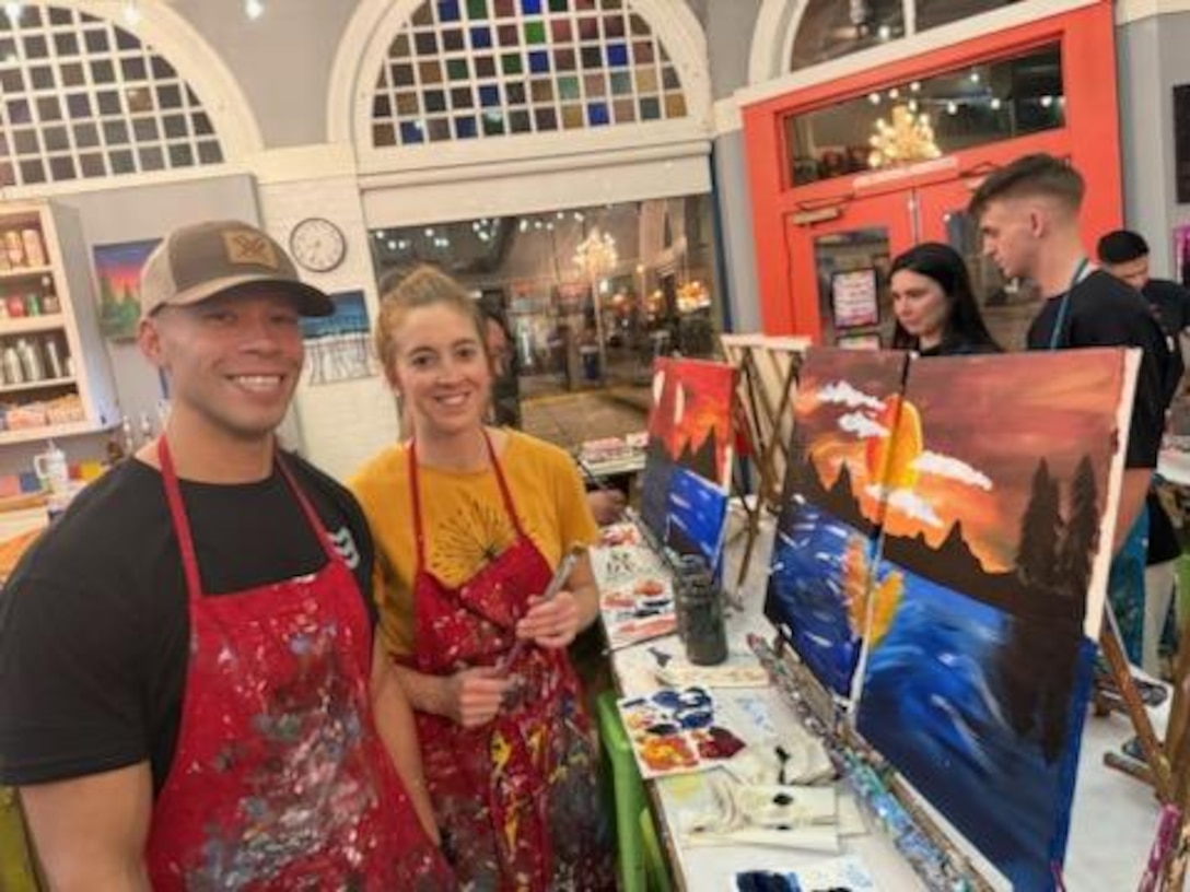 Two people pose and smile in front of canvases painted with a mountain and lake scene in paint