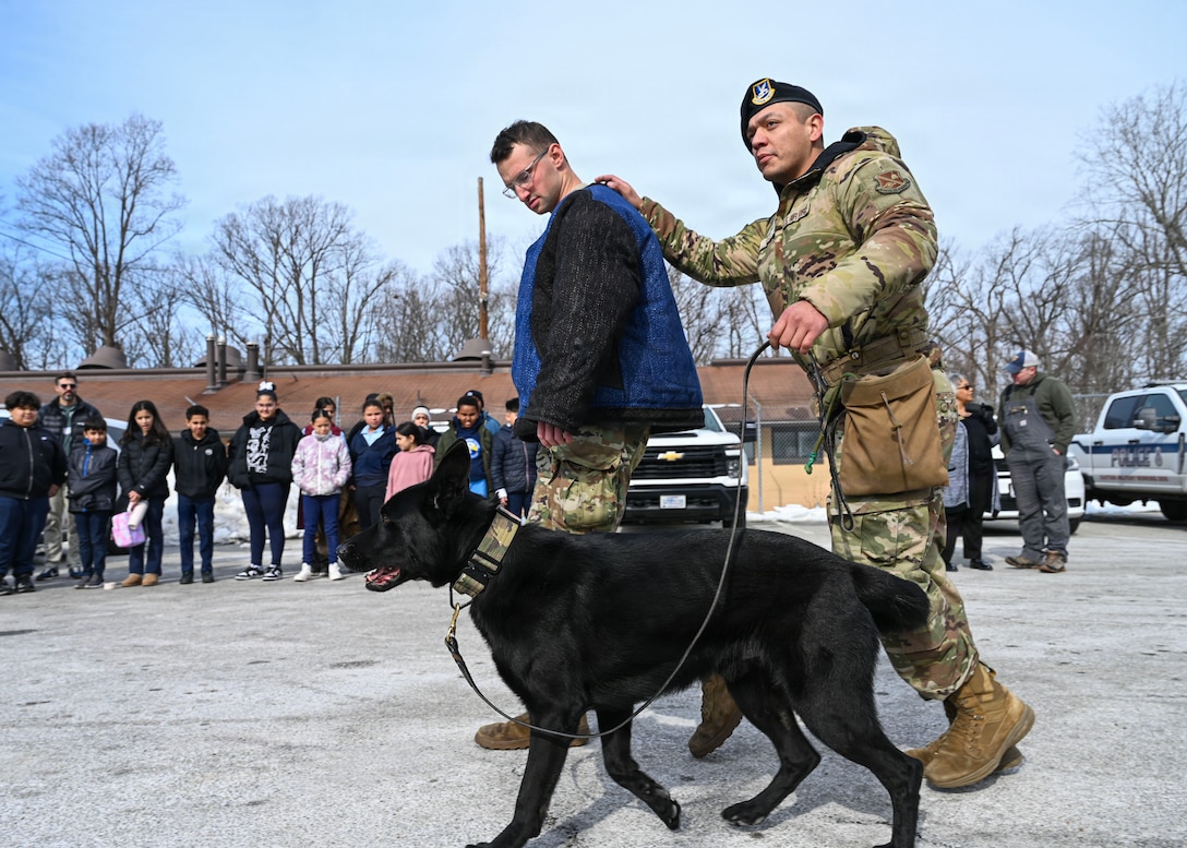 U.S. Air Force Staff Sgt. Alex Reza, 316th Security Support Squadron military working dog handler, right, conducts a military working dog demonstration for students from Patrick Henry K-8 School during a base tour at Joint Base Andrews, Maryland, Feb. 10, 2026. Students were able to visit the MWD flight and 316th Civil Engineer Squadron explosive ordnance disposal flight for a community engagement tour, hosting 46 students from a local school to gain a better understanding of the military. (U.S. Air Force photo by Staff Sgt. Aubree Owens)