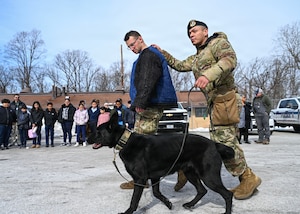 U.S. Air Force Staff Sgt. Alex Reza, 316th Security Support Squadron military working dog handler, right, conducts a military working dog demonstration for students from Patrick Henry K-8 School during a base tour at Joint Base Andrews, Maryland, Feb. 10, 2026. Students were able to visit the MWD flight and 316th Civil Engineer Squadron explosive ordnance disposal flight for a community engagement tour, hosting 46 students from a local school to gain a better understanding of the military. (U.S. Air Force photo by Staff Sgt. Aubree Owens)