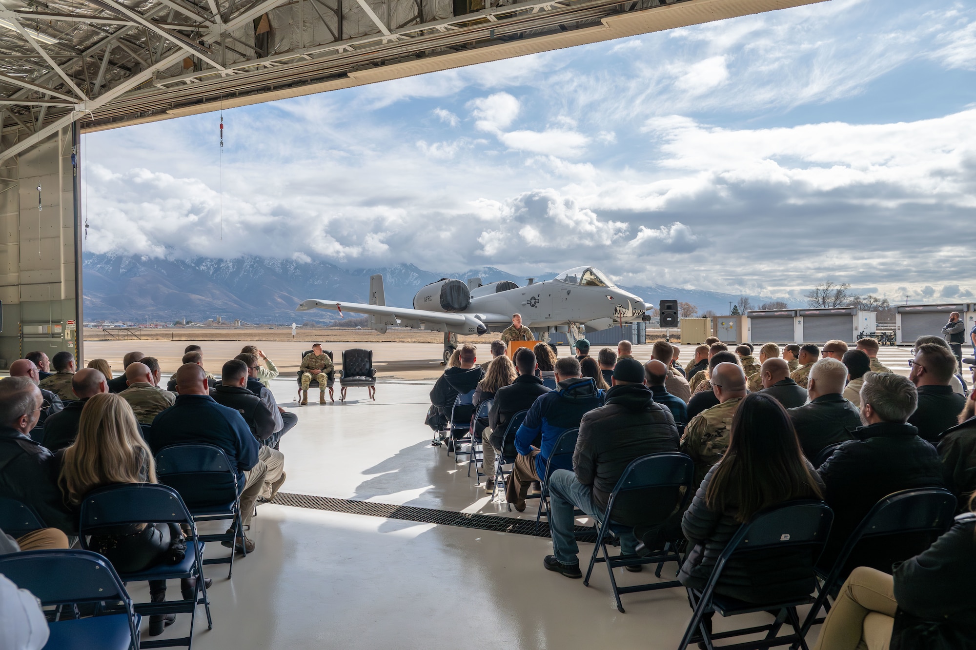 General in military uniform speaks to a crowd while standing in front of an aircraft with painted teeth on the nose