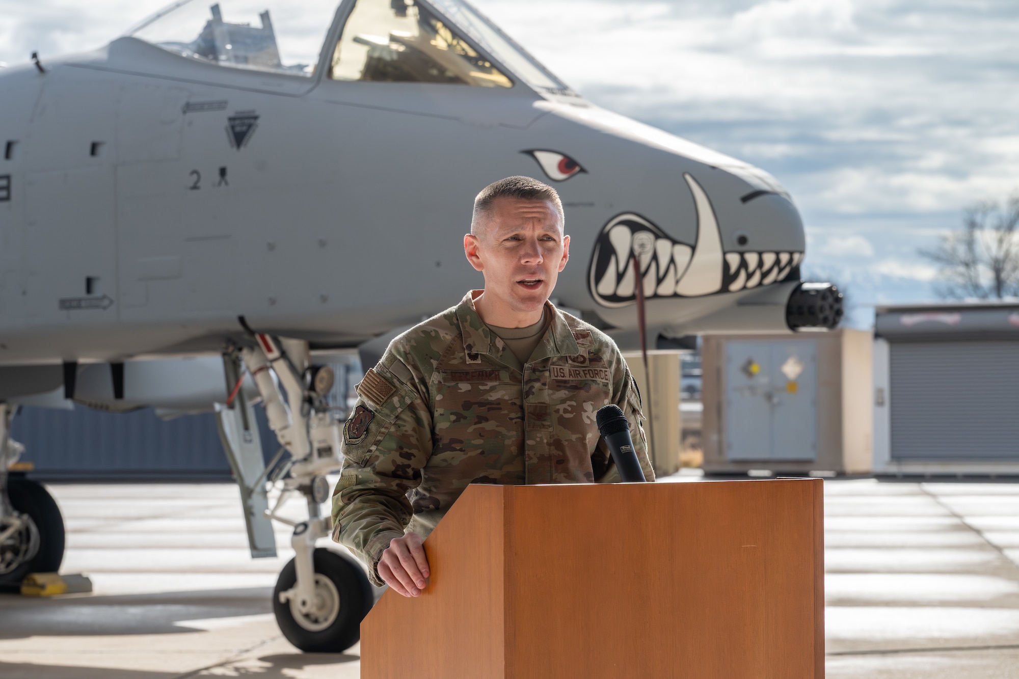 General in military uniform speaks to a crowd while standing in front of an aircraft with painted teeth on the nose