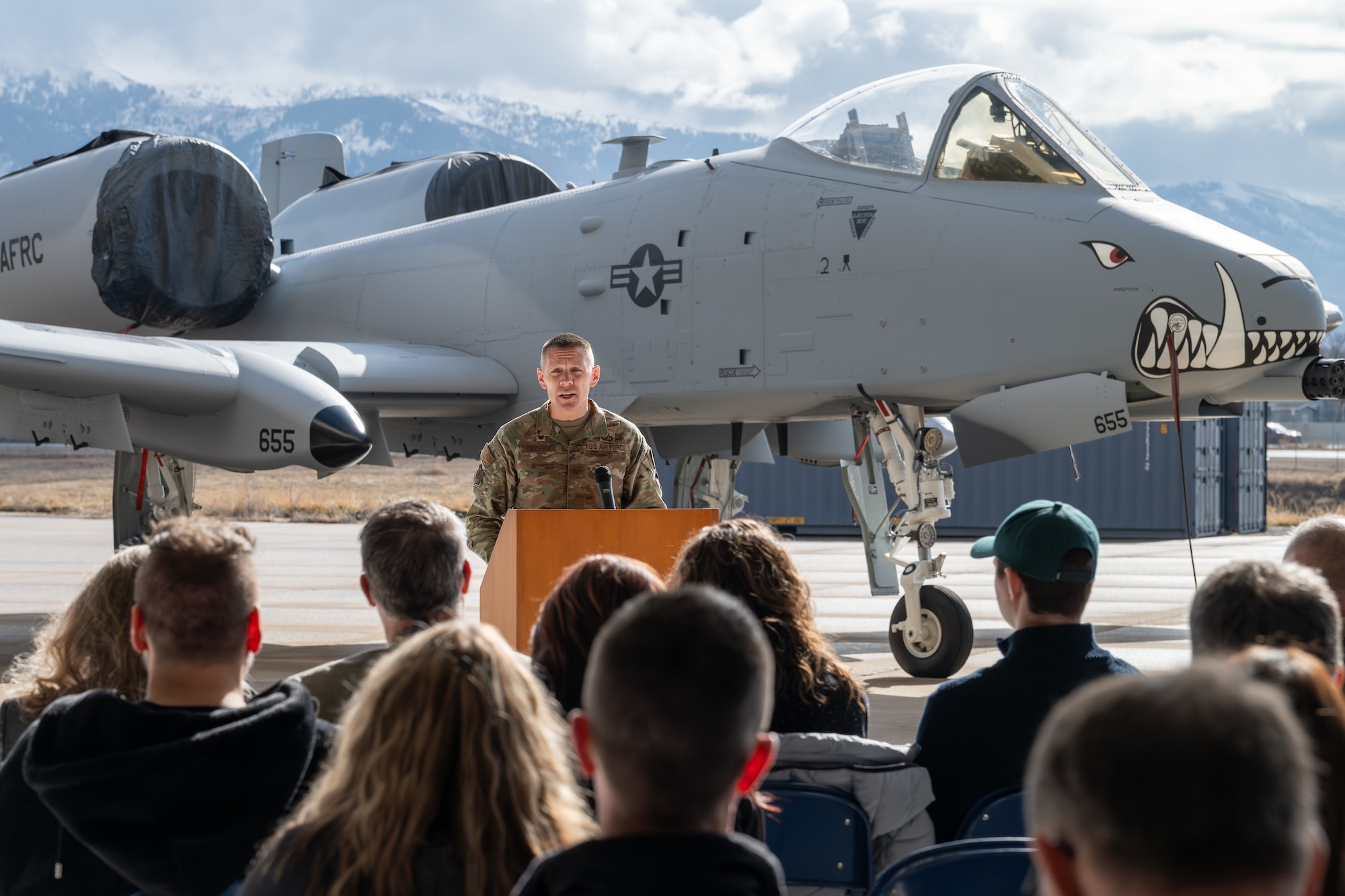 Colonel in military uniform speaks to a crowd while standing in front of an aircraft with painted teeth on the nose