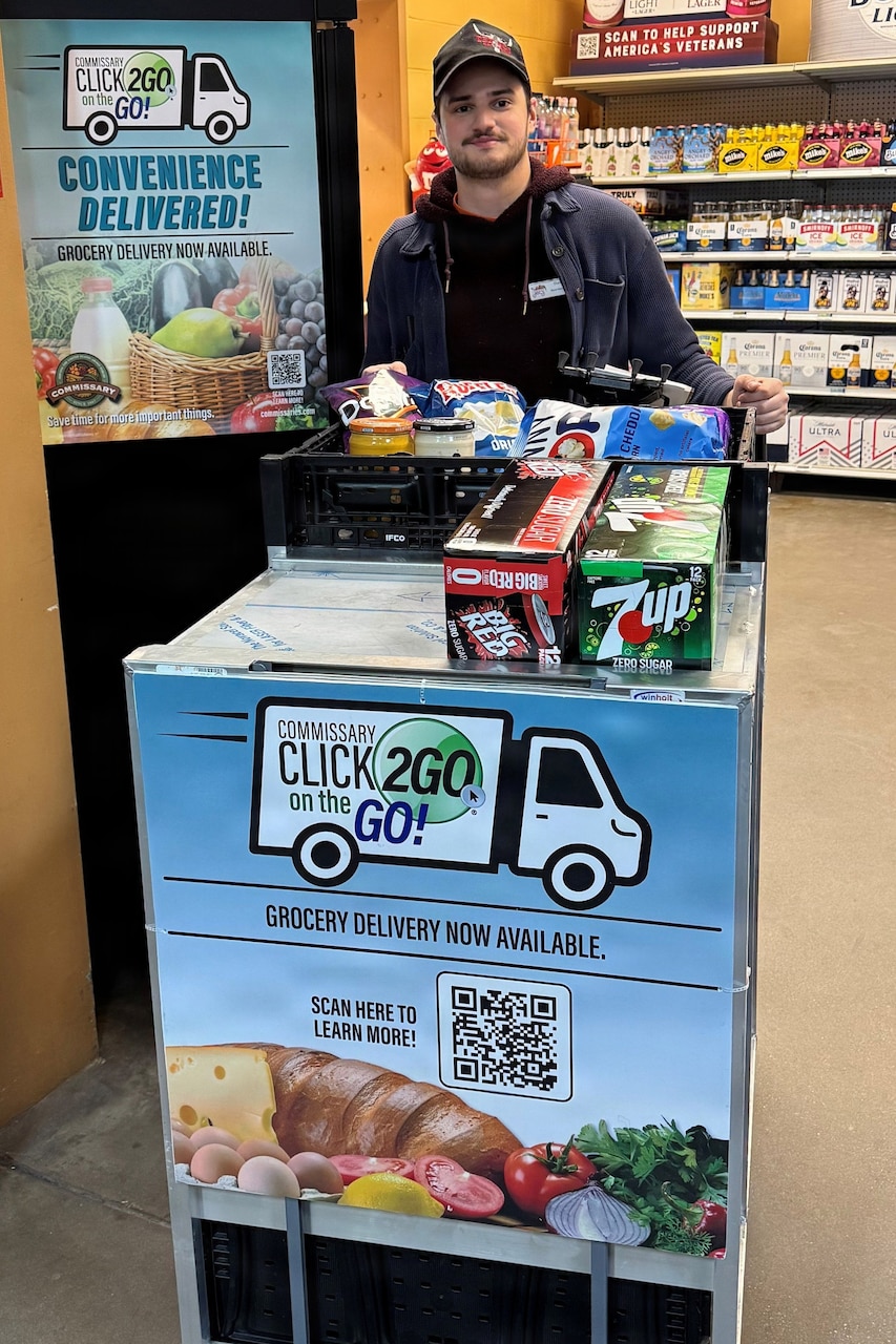 A man in casual attire stands behind a wheeled cart full of drinks and food in a grocery store. The cart has a sign that reads “CLICK2GO® on the GO!"