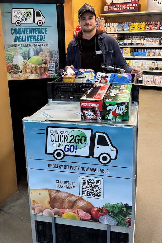 A man in casual attire stands behind a wheeled cart full of drinks and food in a grocery store. The cart has a sign that reads “CLICK2GO® on the GO!"