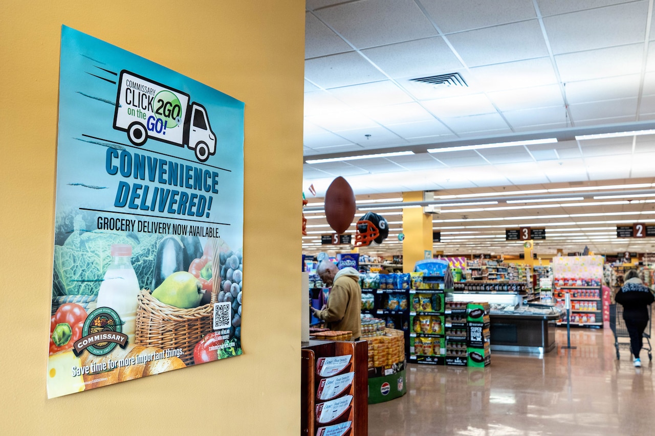 In a grocery store, a sign on the wall reads "CLICK2GO on the GO! Convenience Delivered!" There is a person in casual attire pushing a shopping cart and another person in similar attire standing in the store.