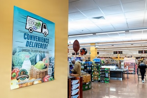 In a grocery store, a sign on the wall reads "CLICK2GO on the GO! Convenience Delivered!" There is a person in casual attire pushing a shopping cart and another person in similar attire standing in the store.