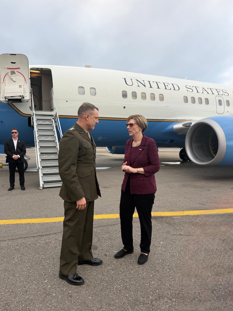 A man in a military uniform speaks with a woman in a purple shirt while standing in front of a large jet.