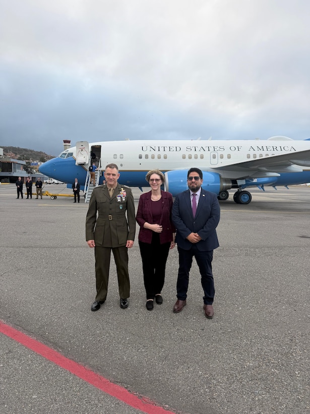 A man in a military uniform, a woman in a purple shirt, and a man in a dark blue suit pose for a photo on a flight line in front of a jet.
