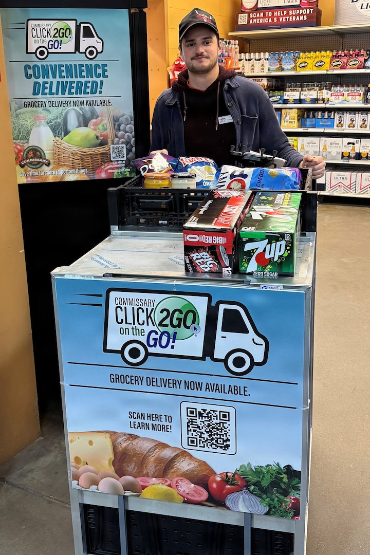 A man in casual attire stands behind a wheeled cart full of drinks and food in a grocery store. The cart has a sign that reads “CLICK2GO® on the GO!"