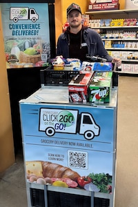A man in casual attire stands behind a wheeled cart full of drinks and food in a grocery store. The cart has a sign that reads “CLICK2GO® on the GO!"