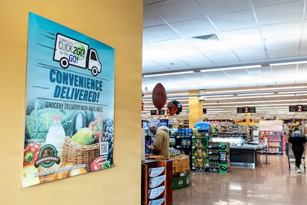 In a grocery store, a sign on the wall reads "CLICK2GO on the GO! Convenience Delivered!" There is a person in casual attire pushing a shopping cart and another person in similar attire standing in the store.
