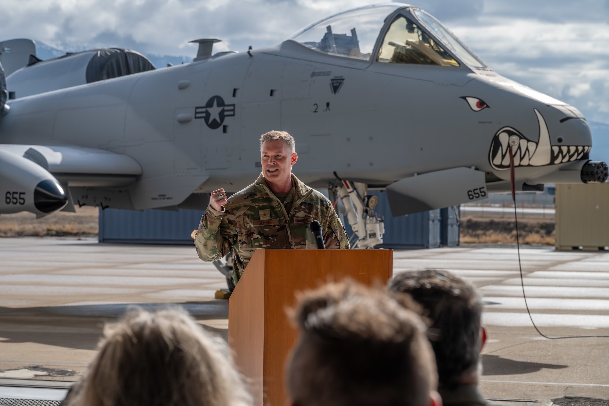 General in military uniform speaks to a crowd while standing in front of an aircraft with painted teeth on the nose