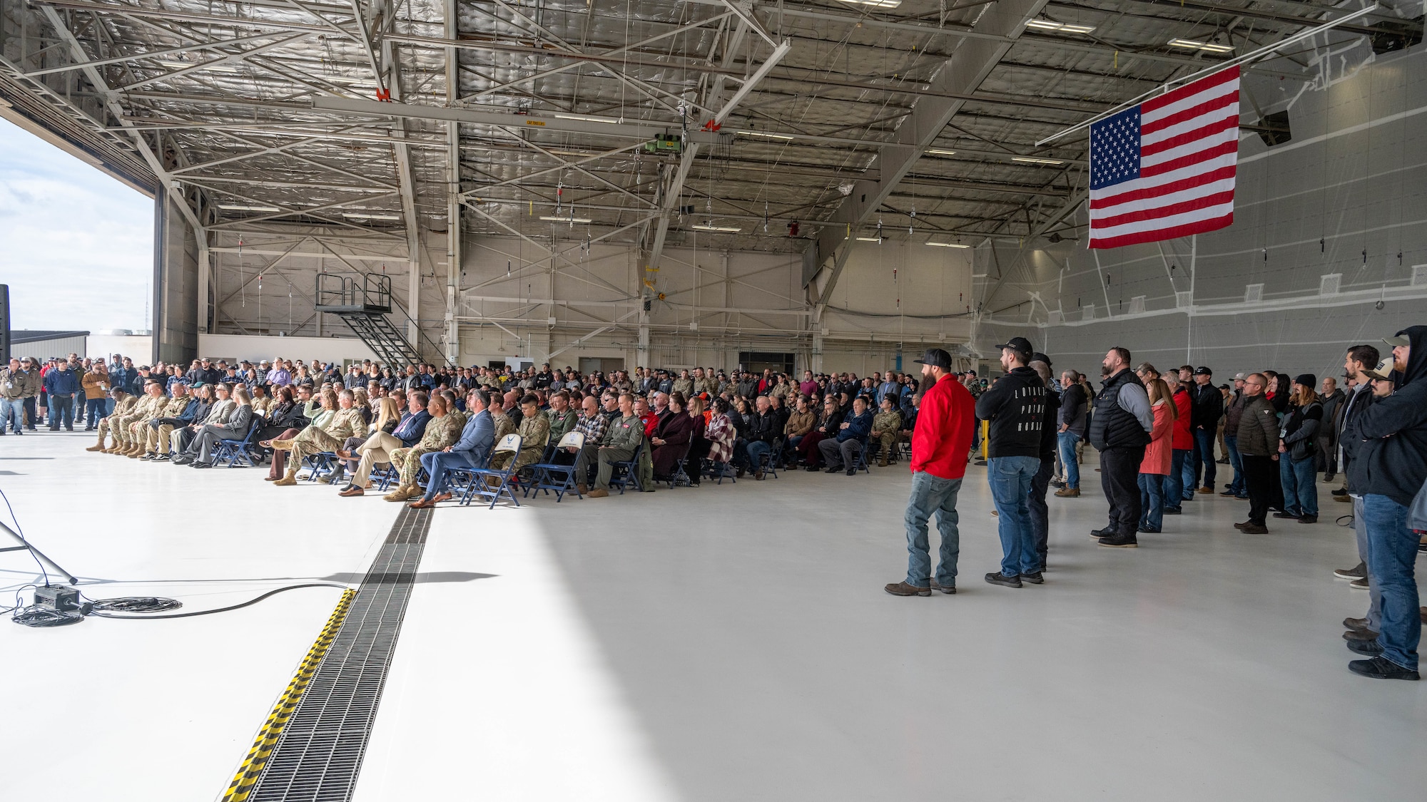 Crowd of many people in an aircraft hangar with US flag hanging from ceiling