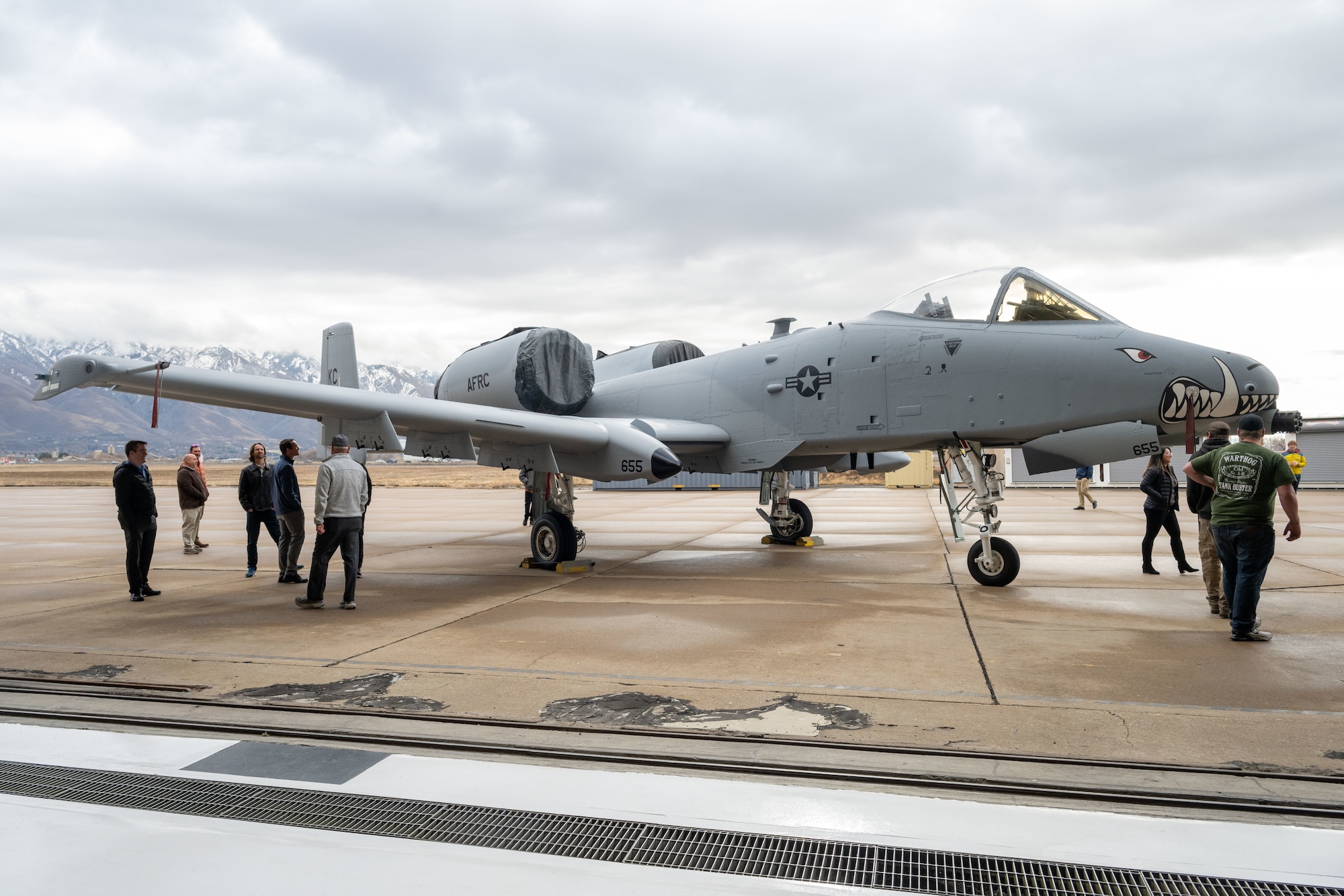 An A10 Aircraft with painted teeth on nose positioned in front of mountains and several people walking around it
