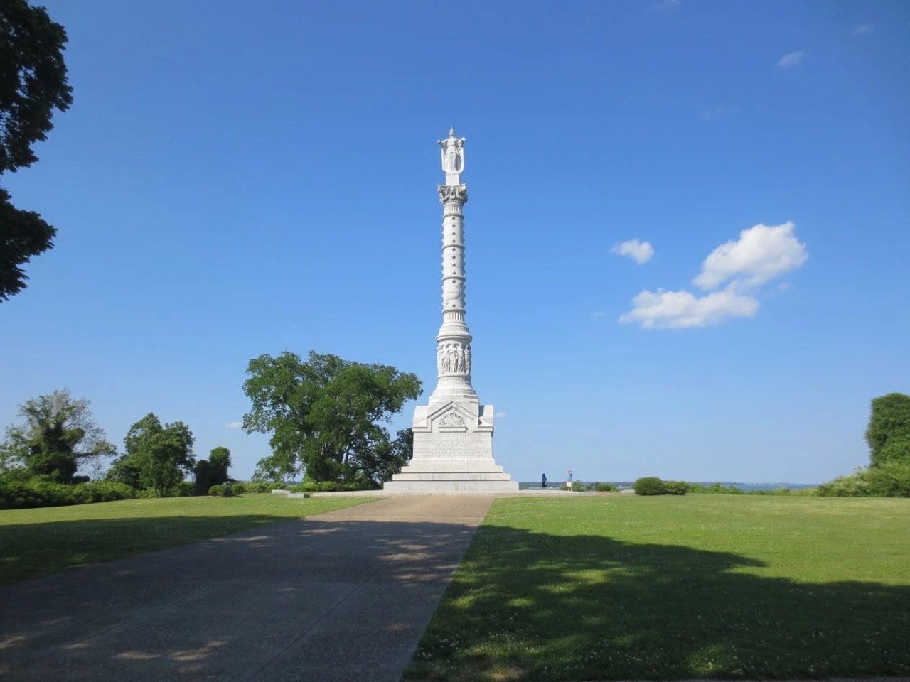 A very tall marble statue topped with a figure at the top rises in a park-like setting under mostly blue skies.