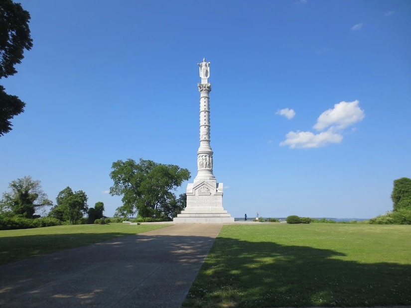 A very tall marble statue topped with a figure at the top rises in a park-like setting under mostly blue skies.