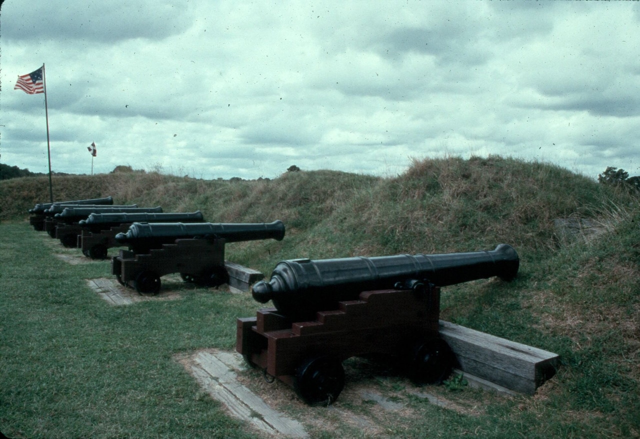 Six cannons on a grassy field point through openings in a berm.