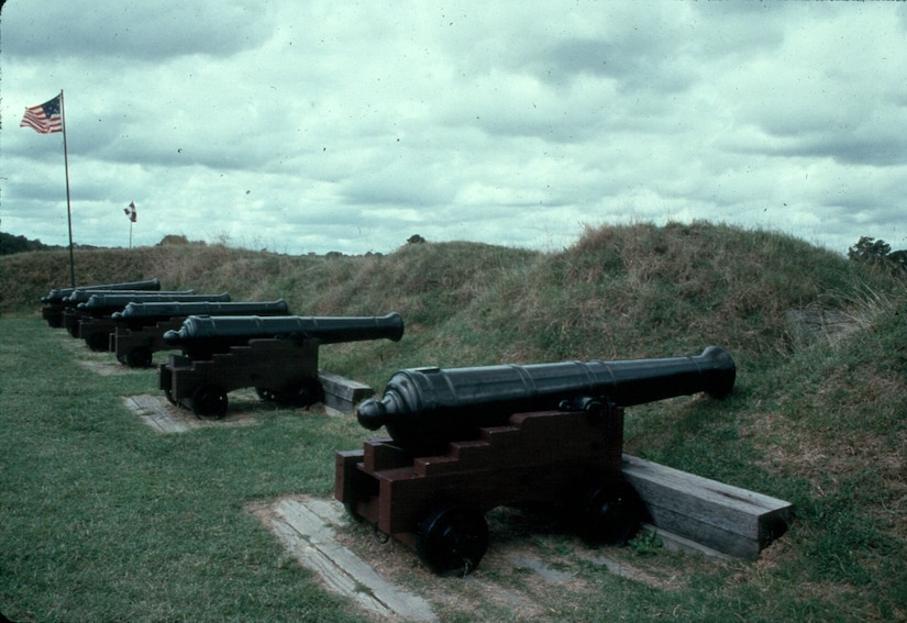 Six cannons on a grassy field point through openings in a berm.