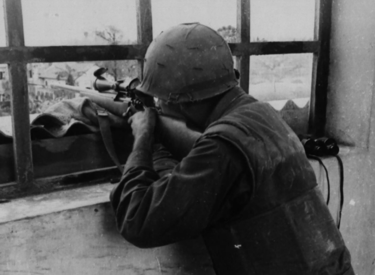 A man wearing a military uniform and a helmet, crouches down and aims a rifle out of the window.