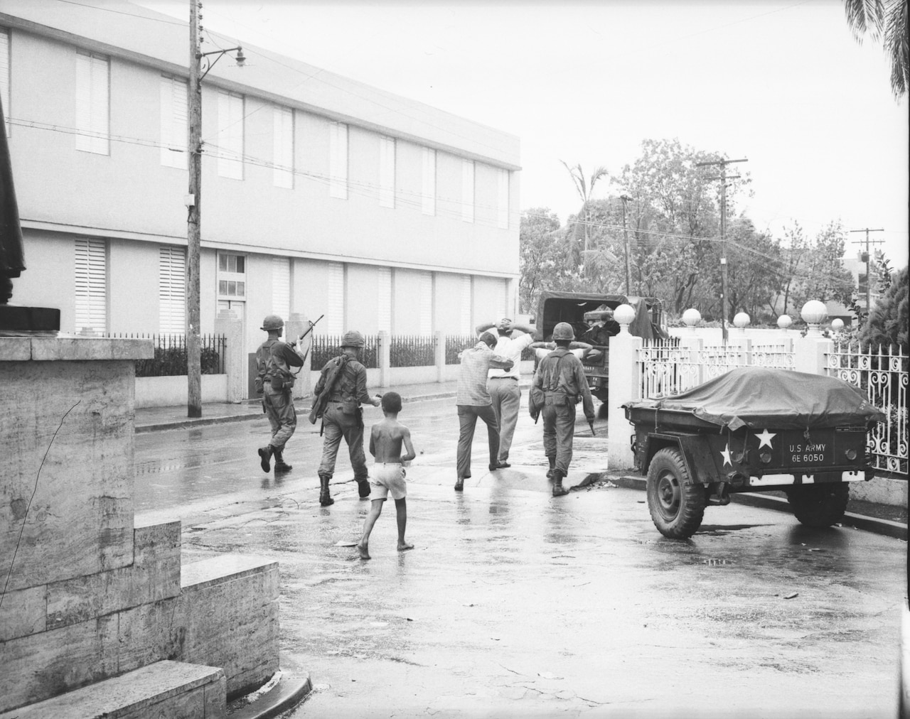 A boy walks behind three people in military uniforms as they usher two men, including one with his hands on his head, toward a military vehicle. They’re walking along an empty street with a building in the background.