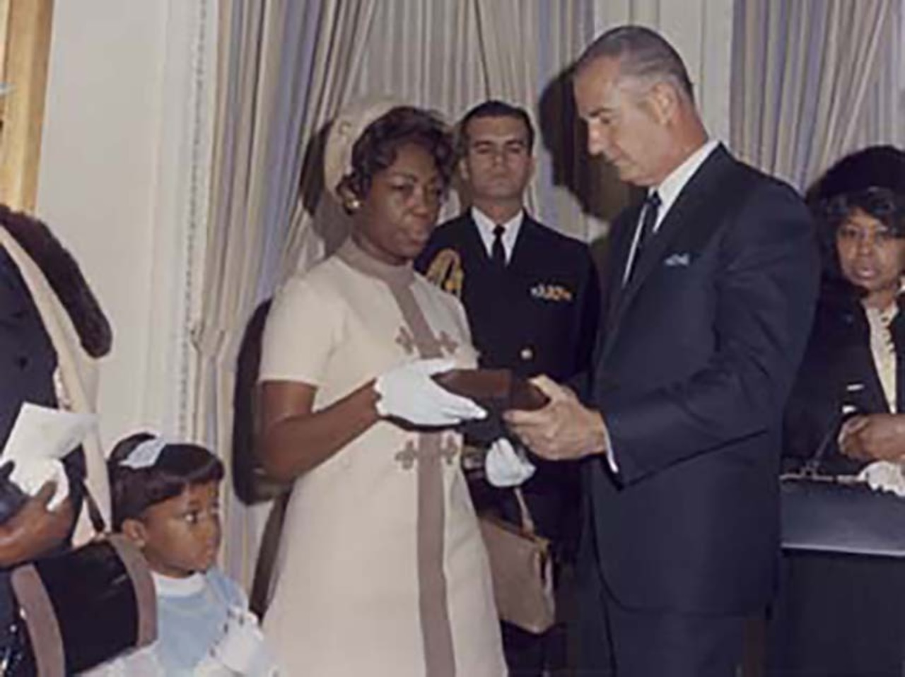 A woman in formal attire is handed a small box from a man in business attire, as people around them, including a little girl, watch.
