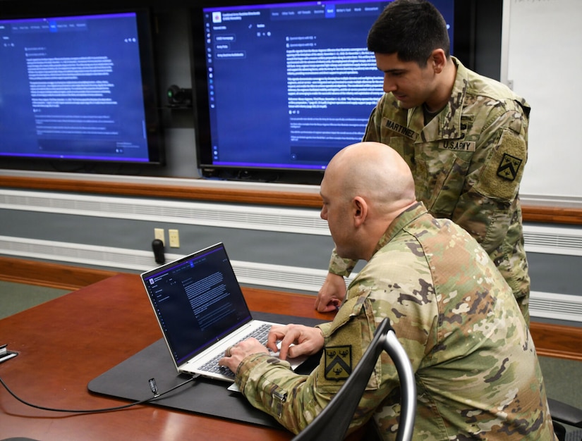 Two men wearing camouflage military uniforms sit at a conference table while looking at a laptop. Two large screens are on a wall next to them.