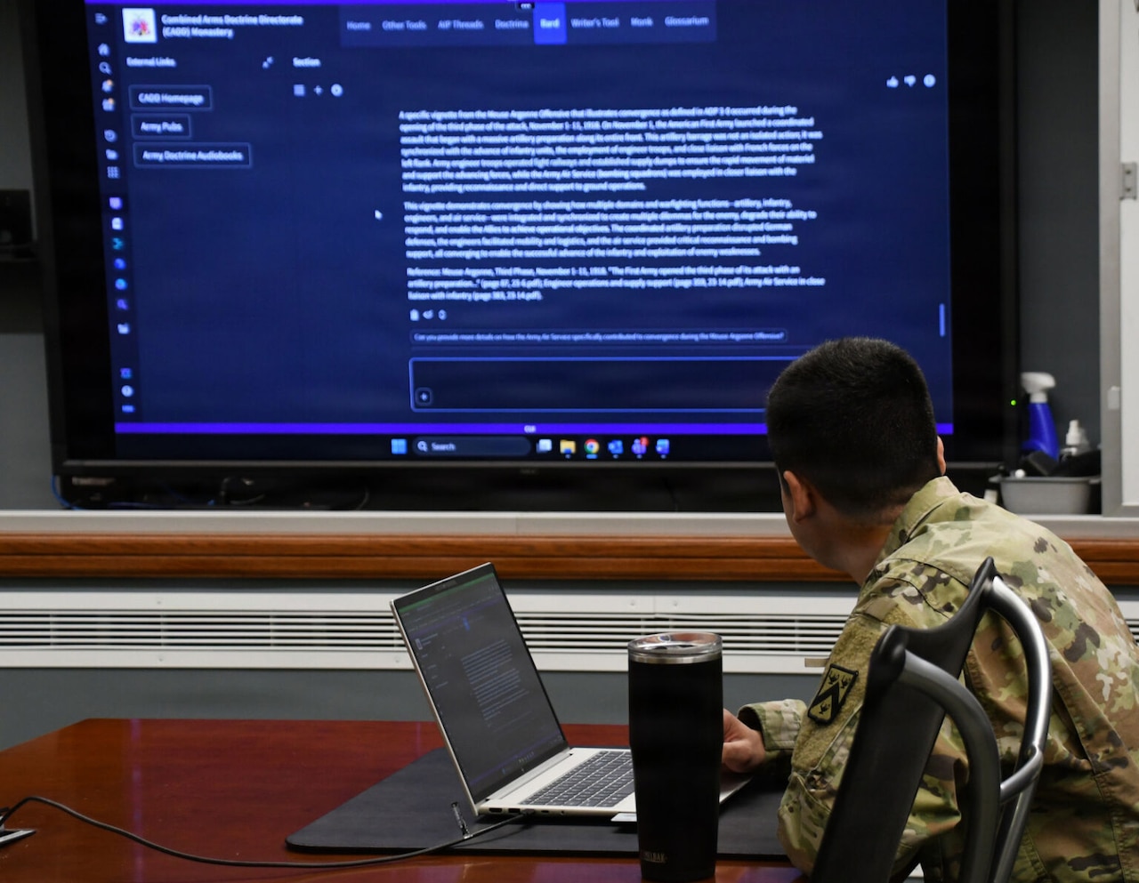 A man wearing a camouflage military uniform sits at a conference table while looking at a large screen on a wall in front of him.