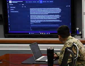 A man wearing a camouflage military uniform sits at a conference table while looking at a large screen on a wall in front of him.