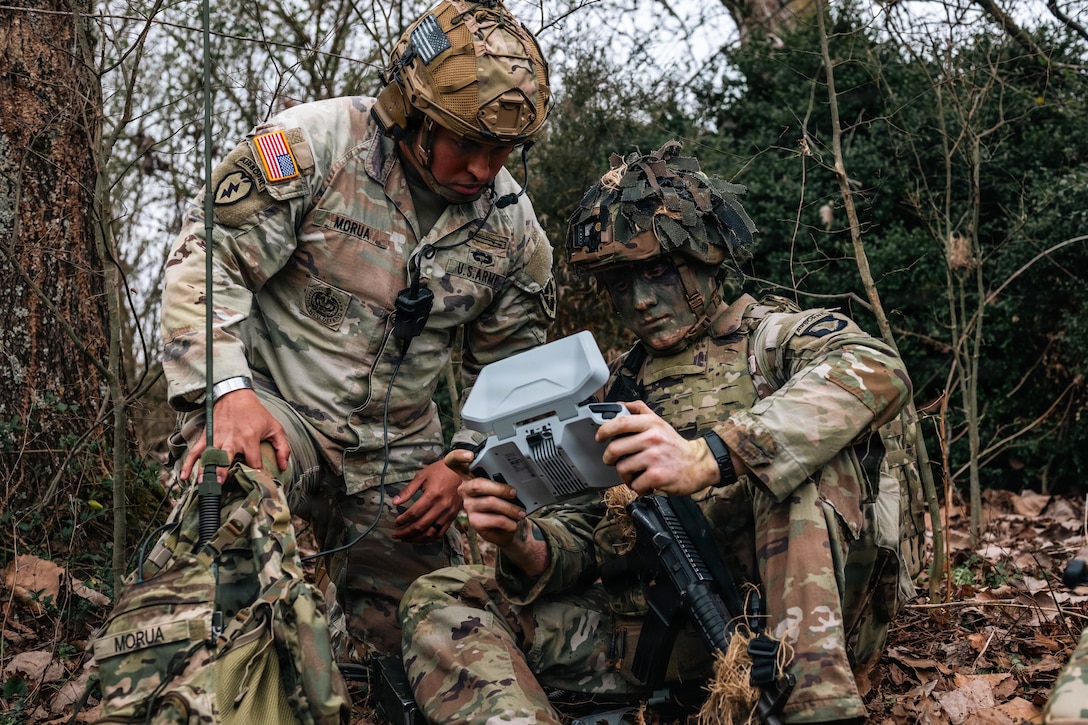 Two people wearing camouflage uniforms and helmets, one of whom has on camouflage face paint, sit in a wooded area and look at an electronic instrument.