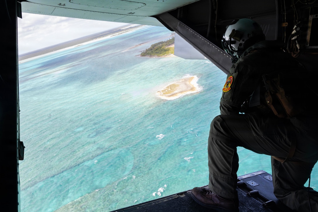 A person wearing a dark green flight suit and helmet kneels in an aircraft doorway, looking out at blue-green water.