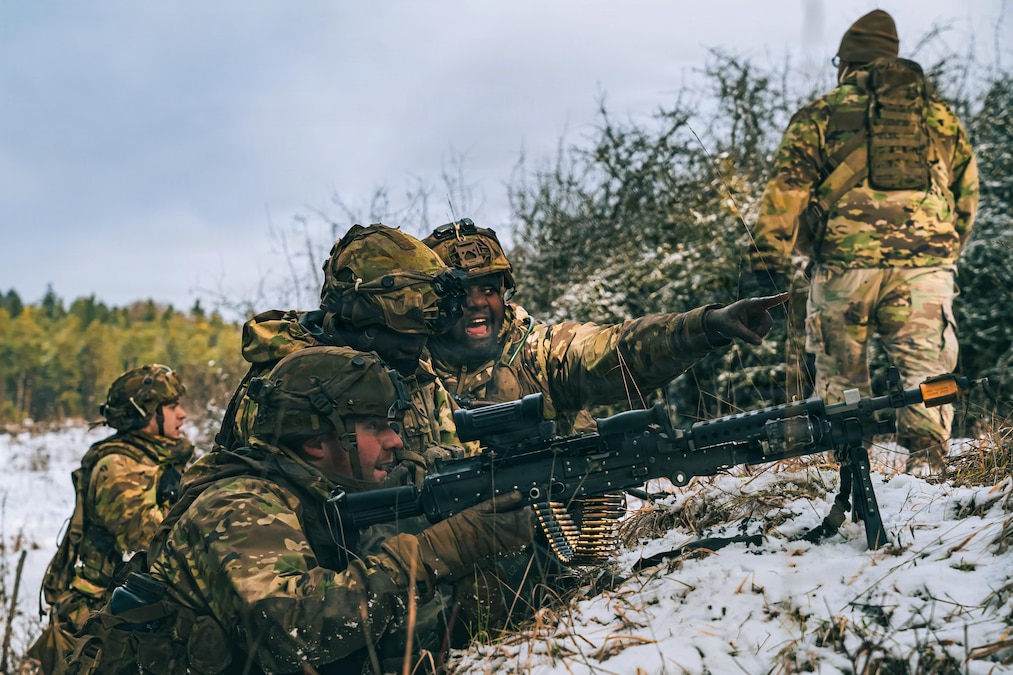 Three people wearing camouflage uniforms and helmets, one lying on his stomach in the snow while holding a weapon and two others kneeling nearby, look toward a target in the distance. Trees and people in similar attire are in the background.