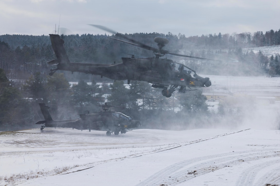 A helicopter lifts off from a snow-covered area, with trees and another helicopter in the background, causing a cloud to whirl around the aircraft.