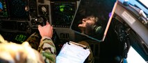 An Airman sits in the seat of a cockpit with their hand on the joystick and a clipboard in her lap. Her face is reflected on a mounted screen in front of her.