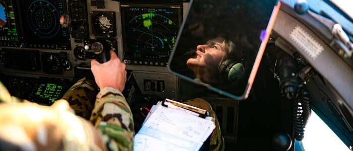 An Airman sits in the seat of a cockpit with their hand on the joystick and a clipboard in her lap. Her face is reflected on a mounted screen in front of her.