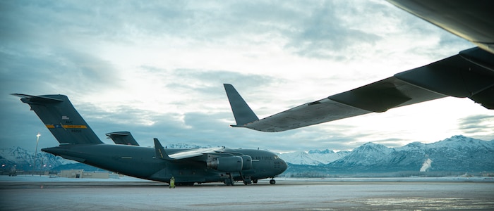 A C-17 aircraft sits parked on the flightline with mountains and a cloudy sky in the background.