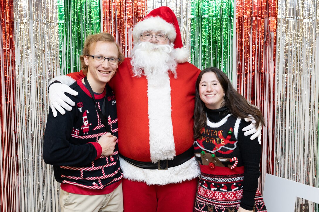 two people in ugly sweater pose with santa clause in front of red and green streamers