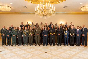 A group of people stand and pose for a photo in a ballroom-like setting, some in military dress uniforms and others in business attire, with a chandelier hanging from the ceiling.