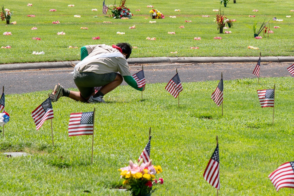 A youth kneels while placing a small U.S. flag at a grave amid rows of similar flags at other graves.